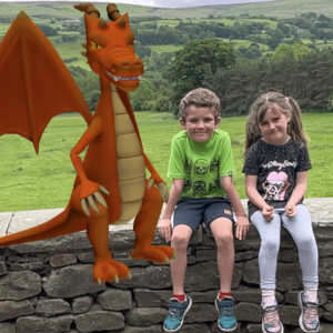 A dragon stands next to a young girl and boy sitting on a stone wall with rolling fields behind them.